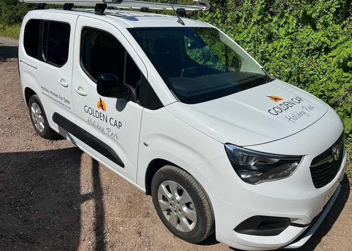 White van parked on a gravel road, featuring a logo on the side. Green foliage in background.