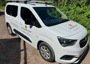 White van parked on a gravel road, featuring a logo on the side. Green foliage in background.