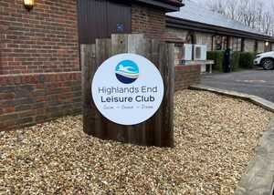 Sign for Highlands End Leisure Club in a gravel area, set against a brick building.