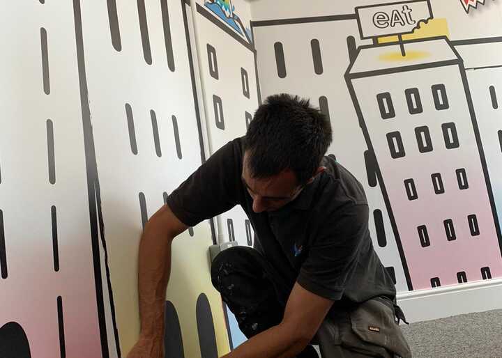 A person kneels while painting colorful building designs on a wall.