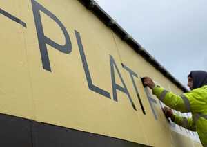 A worker applies letters to a yellow wall, partially revealing the word "PLATFORM."