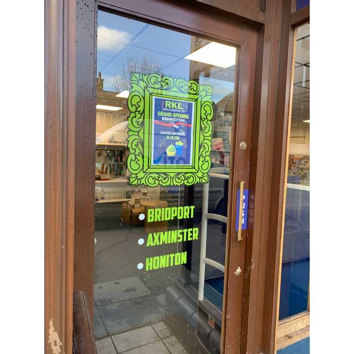 Sign on glass door announcing a grand opening, listing locations: Bridport, Axminster, Honiton.