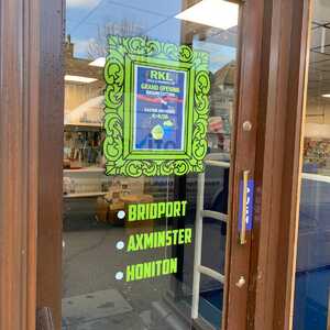 Sign on glass door announcing a grand opening, listing locations: Bridport, Axminster, Honiton.