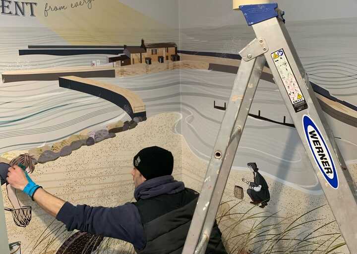 A person applies wallpaper to a wall featuring a seaside mural, using a ladder for support.