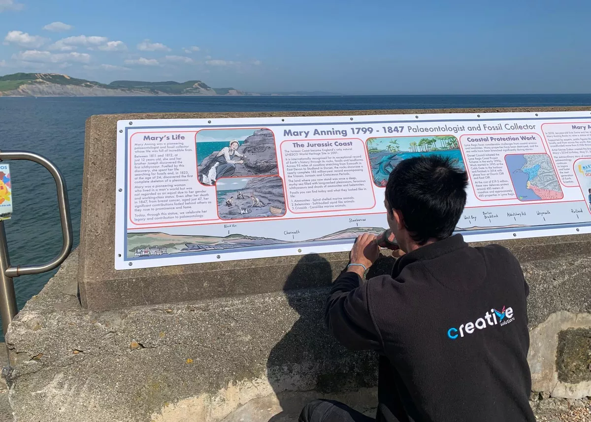 Creative Solutions working on a display board about Mary Anning on the Lyme Regis coast under a blue sky.