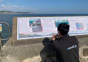 Creative Solutions working on a display board about Mary Anning on the Lyme Regis coast under a blue sky.