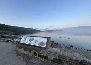 Coastal view with a large format information sign about Mary Anning, calm water, and hills in the background.