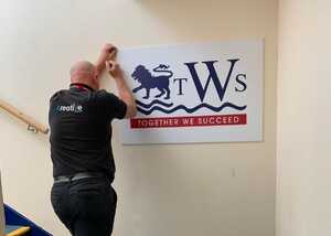 A person hangs a large sign that reads "TWS" with a lion logo on a wall.