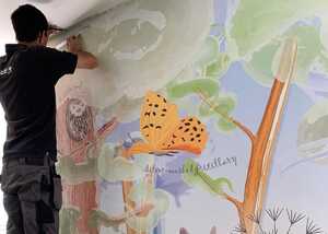 Person painting a colorful mural with trees and a butterfly on a wall.